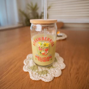 Op de afbeelding: Een heldere glazen beker met een bamboe deksel en rietje, gevuld met een gele drank. Het glas heeft een "STRAWBERRIES PELICAN TOWN" afbeelding. Het staat op een wit en groen gehaakt onderzetter.