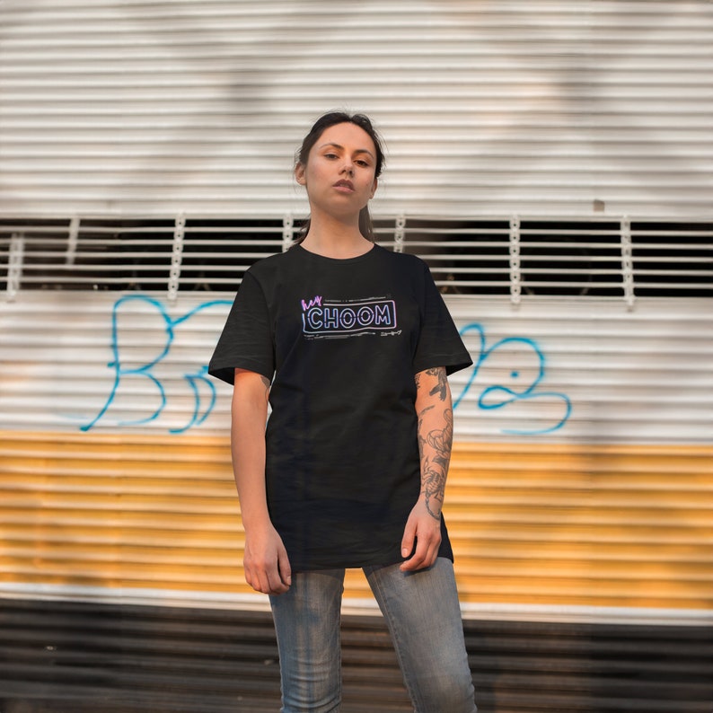 A woman standing in front of a warehouse door covered in graffiti wearing a black Cyberpunk 2077 Hey Choom Unisex Oversized T-Shirt by New Save.