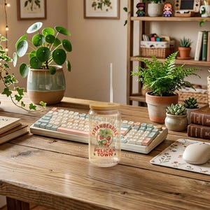 Op de afbeelding: Een heldere glazen beker met een bamboe deksel en een rietje, met een "Strawberries Pelican Town" afbeelding. De beker staat op een houten bureau naast een toetsenbord, muis en potplanten. Het toetsenbord heeft pastelkleurige toetsen.