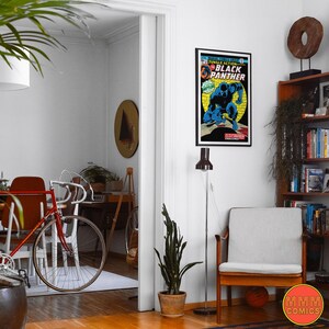 May include: A vintage comic book cover featuring Black Panther in a blue and black costume, with the title "Jungle Action #5" and "Black Panther" in yellow lettering. The comic book is framed and hanging on a white wall in a room with a wooden floor, a red bicycle, and a potted plant.