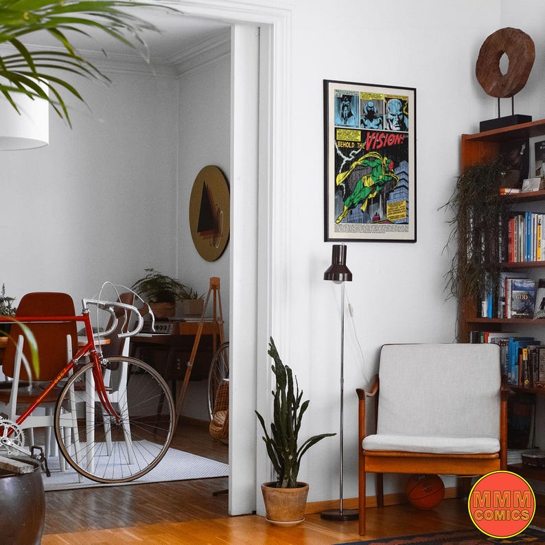 May include: Interior shot of a living room with a red bicycle, a framed comic book poster, and a wooden bookshelf. A white armchair and a floor lamp are also visible. The room has a minimalist design with neutral colors.