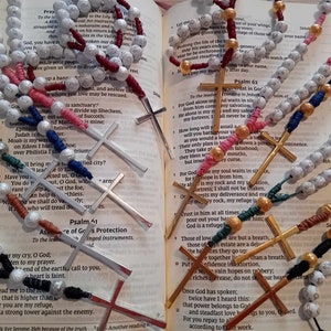 Puede incluir: Una colección de rosarios de cuentas de colores con cruces de plata y oro, colocados sobre una Biblia abierta con texto de los Salmos.