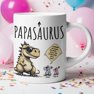 Puede incluir: Taza de cer&aacute;mica blanca con la inscripci&oacute;n "Papasaurus" y un dinosaurio de dibujos animados con gafas de sol. La taza tambi&eacute;n presenta dos personajes de dinosaurios m&aacute;s peque&ntilde;os y bocadillos de texto con texto en franc&eacute;s. Globos de colores en el fondo.
