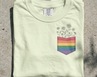 May include: A pale yellow t-shirt featuring a graphic of a chest pocket with horizontal rainbow stripes. Above the pocket, a line drawing of several daisies and wildflowers extends upward. The inner neck label reads Comfort Colors.