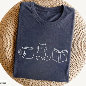 May include: Dark heather gray t-shirt with a white line drawing of a teacup with a heart, a cat, and an open book with a heart. The text "dark heather" is at the bottom left of the image. The shirt is folded on a woven surface.