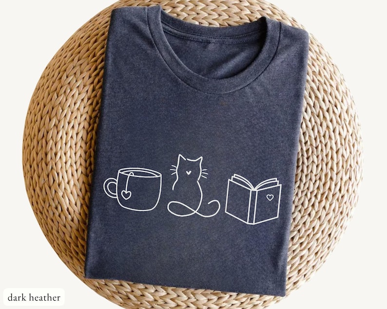 May include: Dark heather gray t-shirt with a white line drawing of a teacup with a heart, a cat, and an open book with a heart. The text "dark heather" is at the bottom left of the image. The shirt is folded on a woven surface.