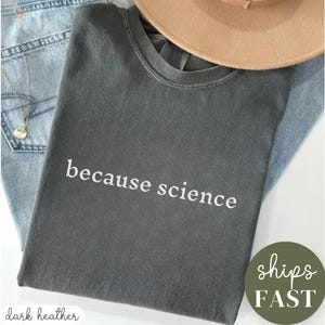 May include: Dark heather gray t-shirt with the text "because science" in white. The shirt is folded on top of blue jeans and a tan hat. A green circle with the words "ships FAST" is in the lower right corner.