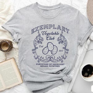 Könnte beinhalten: Hellgraues T-Shirt mit einem marineblauen Aufdruck, der „Exemplary Vegetable Club“ mit einer Illustration von drei Kartoffeln zeigt. Der Text „Purveyors of Excellent Boiled Potatoes“ steht darunter. Das T-Shirt ist am unteren Ende geknotet.