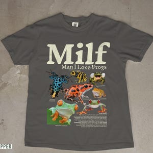 May include: Pepper-colored t-shirt featuring the text "Milf Man I Love Frogs" in white. The shirt displays colorful illustrations of various frog species. A fun and quirky design for frog enthusiasts.