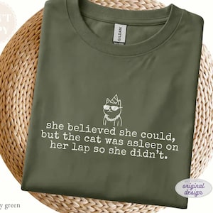 May include: A military green crew neck t-shirt featuring a white line art graphic of a cat wearing sunglasses, a party hat, and a chain. The text reads: she believed she could, but the cat was asleep on her lap so she didn't. The shirt has a Gildan label.