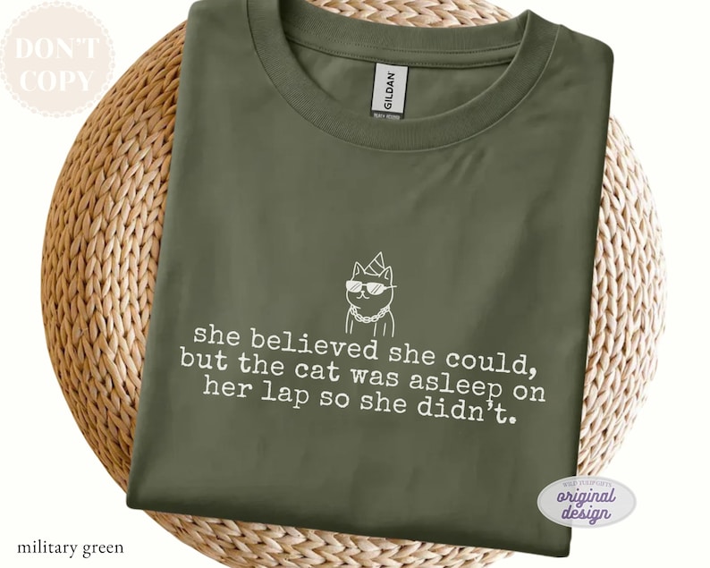 May include: A military green crew neck t-shirt featuring a white line art graphic of a cat wearing sunglasses, a party hat, and a chain. The text reads: she believed she could, but the cat was asleep on her lap so she didn't. The shirt has a Gildan label.