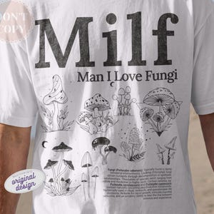 May include: White t-shirt with the text "Milf, Man I Love Fungi" in black block letters. Below the text are several black and white illustrations of different types of mushrooms. The shirt has a round neck and short sleeves.