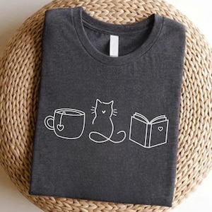 Peut inclure: T-shirt gris foncé avec un motif blanc représentant une tasse à thé, un chat et un livre. La tasse à thé a un cœur, le chat est assis et le livre a un cœur. Le t-shirt est plié sur une surface circulaire tissée.