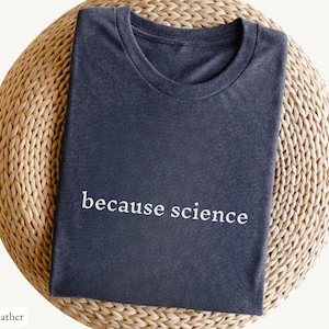 Op de afbeelding: Donkergrijs heather t-shirt met de witte tekst "because science", gepresenteerd op een geweven, ronde ondergrond. Het shirt heeft een ronde hals en is netjes opgevouwen. De stijl is casual.