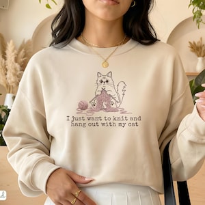 May include: Sand-colored sweatshirt with a graphic of a cat knitting. The cat is depicted with yarn and knitting needles, and the text "I just want to knit and hang out with my cat" is printed below the graphic.