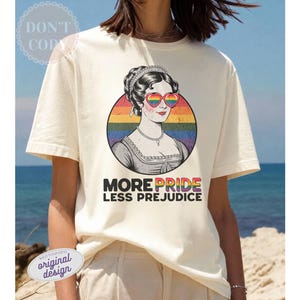 Puede incluir: Camiseta color crema con una ilustración vintage de una mujer con gafas de sol en forma de corazón arcoíris. La imagen está dentro de un círculo arcoíris, con el texto "MORE PRIDE LESS PREJUDICE" debajo. La camiseta tiene mangas cortas.