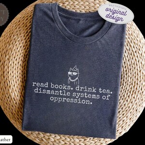 May include: A dark heather gray crew neck t-shirt featuring a white line art graphic of a cat wearing sunglasses and a party hat, with the text "read books. drink tea. dismantle systems of oppression." printed in a typewriter-style font below.