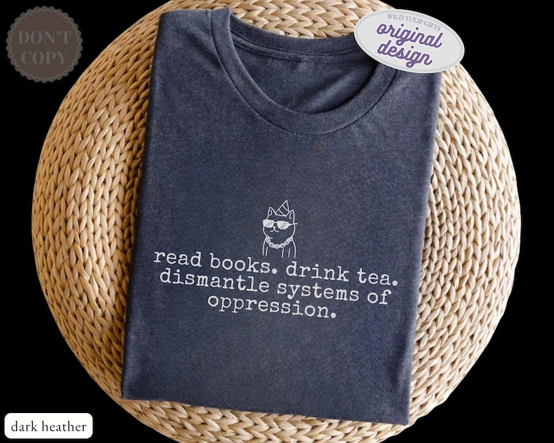 May include: A dark heather gray crew neck t-shirt featuring a white line art graphic of a cat wearing sunglasses and a party hat, with the text "read books. drink tea. dismantle systems of oppression." printed in a typewriter-style font below.