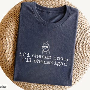 May include: Dark heather gray t-shirt with the text "if i shenan once, i'll shenanigan" and a cat wearing sunglasses and a crown. The shirt is folded on a woven, circular surface.