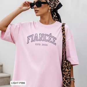 May include: Light pink t-shirt with the word "FIANC&Eacute;E" in navy blue block letters and "ESTD. 2026" underneath. The shirt has short sleeves and a relaxed fit. A leopard print tote bag is visible.