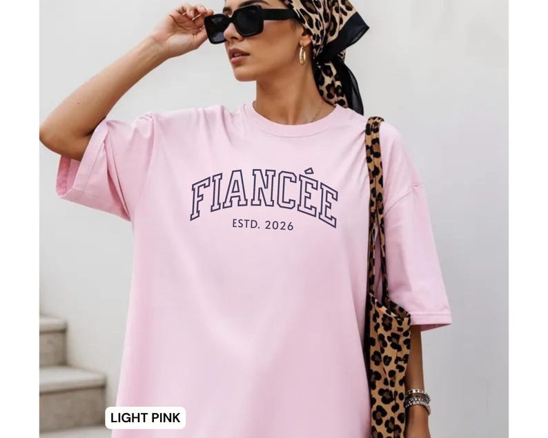 May include: Light pink t-shirt with the word "FIANC&Eacute;E" in navy blue block letters and "ESTD. 2026" underneath. The shirt has short sleeves and a relaxed fit. A leopard print tote bag is visible.