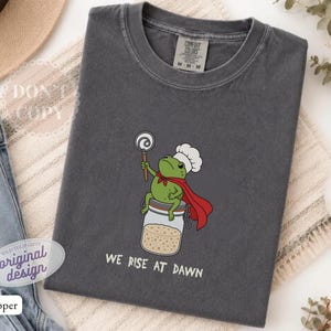 Op de afbeelding: Een antracietgrijs t-shirt met een afbeelding van een groene kikker met een witte koksmuts en een rode cape, zittend op een glazen pot terwijl hij een garde vasthoudt. De tekst "WE RISE AT DAWN" staat in het wit onder de kikker afgebeeld.