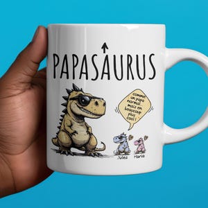 Puede incluir: Taza de cer&aacute;mica blanca con la palabra "PAPASAURUS" y una ilustraci&oacute;n de un dinosaurio con gafas de sol. La taza tambi&eacute;n presenta dos dinosaurios m&aacute;s peque&ntilde;os y texto en un bocadillo.