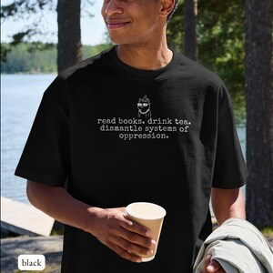 May include: A black short-sleeve t-shirt featuring a white line drawing of a cat wearing glasses and a party hat, with the text "read books. drink tea. dismantle systems of oppression." printed in a typewriter-style font on the chest.
