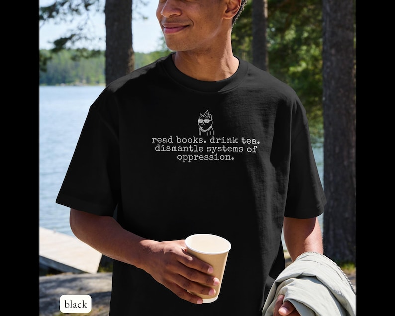 May include: A black short-sleeve t-shirt featuring a white line drawing of a cat wearing glasses and a party hat, with the text "read books. drink tea. dismantle systems of oppression." printed in a typewriter-style font on the chest.