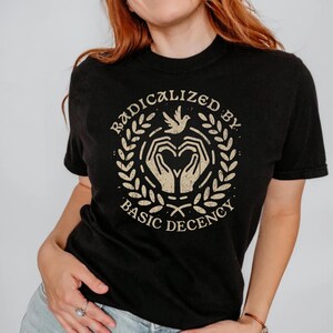 May include: Black t-shirt with the text "Radicalized by Basic Decency" in a circular design. The graphic includes hands in a heart shape, a dove, and a laurel wreath, all in a vintage style.