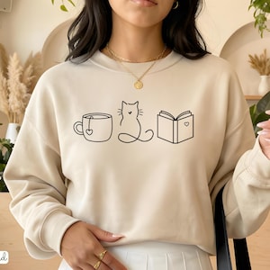 May include: Sand-colored sweatshirt featuring a minimalist black line drawing of a teacup, a cat, and an open book with a heart. The word "sand" is printed in the bottom left corner. The sweatshirt has a crew neck and long sleeves.