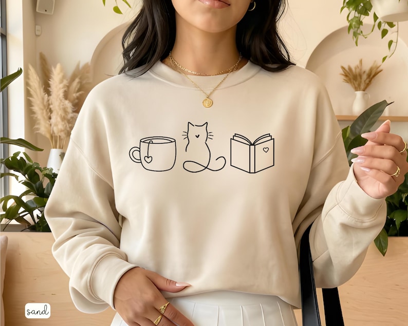 May include: Sand-colored sweatshirt featuring a minimalist black line drawing of a teacup, a cat, and an open book with a heart. The word "sand" is printed in the bottom left corner. The sweatshirt has a crew neck and long sleeves.