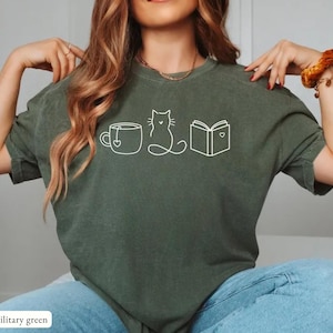 May include: Military green t-shirt with a white outline design of a teacup, cat, and book. The book and teacup have a heart design. The shirt is made of a soft material.