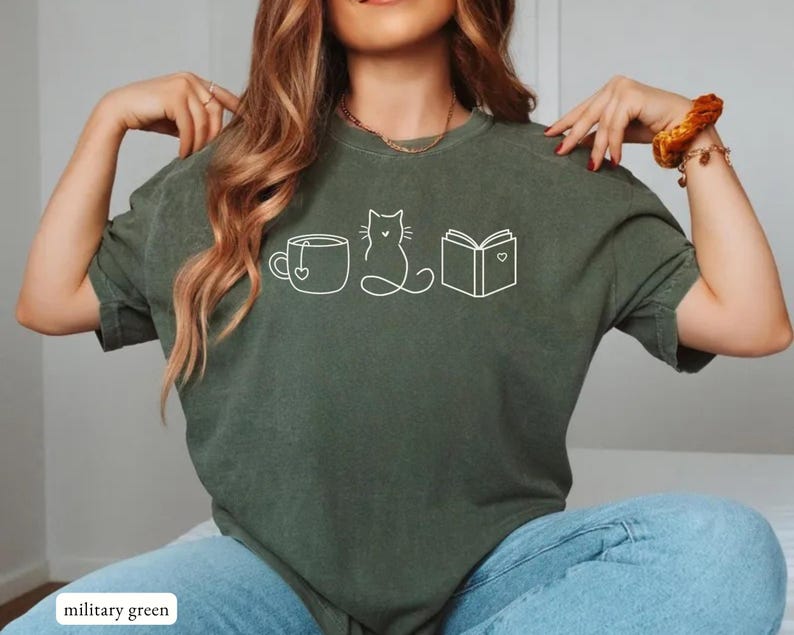 May include: Military green t-shirt with a white outline design of a teacup, cat, and book. The book and teacup have a heart design. The shirt is made of a soft material.