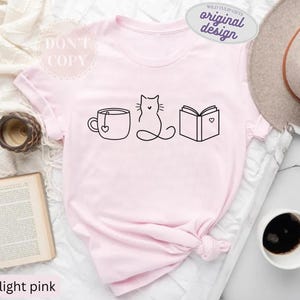 May include: Light pink t-shirt with a black line drawing of a teacup, cat, and book. The teacup has a heart-shaped tag. The book has a heart on it. The shirt also has the text "DON'T COPY" and "original design".