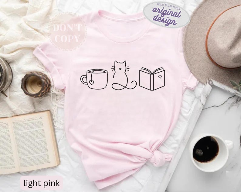 May include: Light pink t-shirt with a black line drawing of a teacup, cat, and book. The teacup has a heart-shaped tag. The book has a heart on it. The shirt also has the text "DON'T COPY" and "original design".