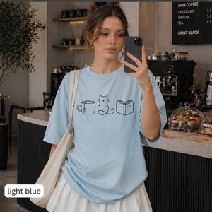 May include: Light blue t-shirt with a black line drawing of a coffee cup, cat, and book. The shirt has short sleeves and a round neckline. The words "light blue" are in a white rectangle at the bottom left.