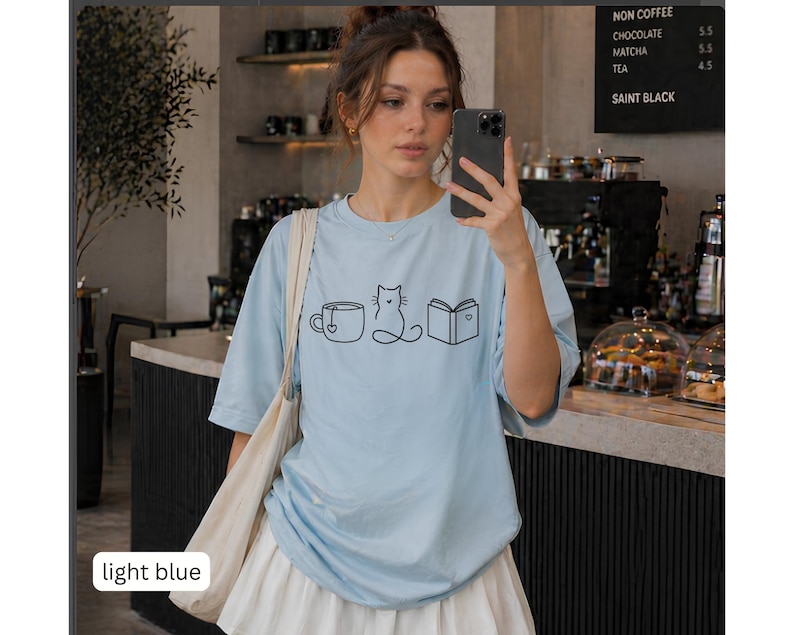 May include: Light blue t-shirt with a black line drawing of a coffee cup, cat, and book. The shirt has short sleeves and a round neckline. The words "light blue" are in a white rectangle at the bottom left.