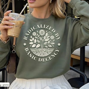 Può includere: Felpa verde militare con la scritta "RADICALIZED BY BASIC DECENCY" in un design circolare. Il design presenta un albero con radici, foglie e fiori. La felpa è a tinta unita e ha maniche lunghe.
