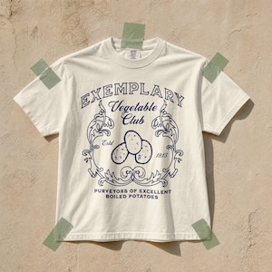 Może przedstawiać: Kremowy t-shirt z krótkim rękawem i ciemnoniebieskim nadrukiem graficznym. Wzór zawiera tekst "EXEMPLARY Vegetable Club", "Estd 1813" oraz "PURVEYORS OF EXCELLENT BOILED POTATOES" otaczający ilustrację przedstawiającą trzy ziemniaki.
