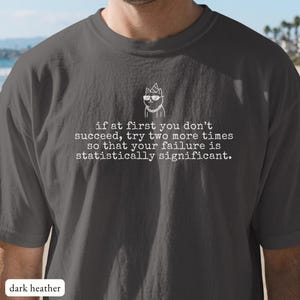 May include: A dark heather grey t-shirt featuring a white line drawing of a cat wearing sunglasses and a party hat. Below the cat, the text reads: if at first you don't succeed, try two more times so that your failure is statistically significant.