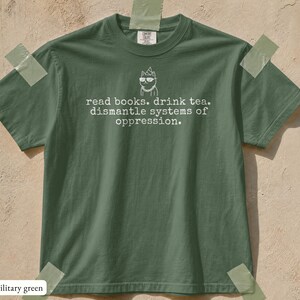 May include: A military green short-sleeve t-shirt featuring a white line-art graphic of a cat wearing sunglasses and a party hat. Below the cat, the text reads: read books. drink tea. dismantle systems of oppression. The shirt has a crew neckline.