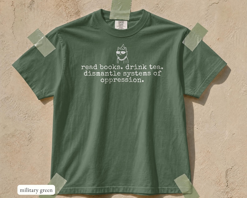 May include: A military green short-sleeve t-shirt featuring a white line-art graphic of a cat wearing sunglasses and a party hat. Below the cat, the text reads: read books. drink tea. dismantle systems of oppression. The shirt has a crew neckline.