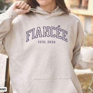 May include: Sand-colored hoodie with the word "FIANC&Eacute;E" in navy blue collegiate lettering above "ESTD. 2026". The hoodie has a drawstring hood and a front pocket. A white mug with a spoon is held in the right hand.