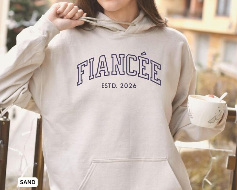 May include: Sand-colored hoodie with the word "FIANC&Eacute;E" in navy blue collegiate lettering above "ESTD. 2026". The hoodie has a drawstring hood and a front pocket. A white mug with a spoon is held in the right hand.