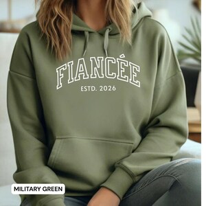 May include: Military green hoodie with the word "FIANC&Eacute;E" in white, arched across the chest, with "ESTD. 2026" below. The hoodie has a drawstring hood and a front pocket.