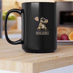 Puede incluir: Taza de cer&aacute;mica negra con un dinosaurio de dibujos animados con gafas de sol y un globo en forma de coraz&oacute;n. El texto "PAPASAURUS" est&aacute; impreso debajo en color dorado. La taza tiene un asa grande y curvada.