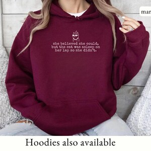 May include: A maroon long-sleeve hooded sweatshirt featuring a white line-art graphic of a cat wearing sunglasses and a party hat, with the text: she believed she could, but the cat was asleep on her lap so she didn't. The hoodie has a front pocket.