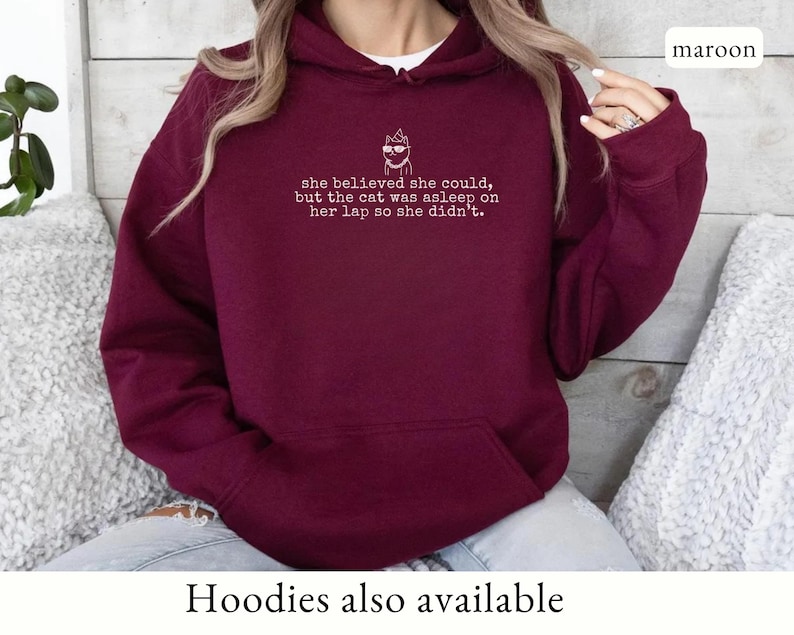 May include: A maroon long-sleeve hooded sweatshirt featuring a white line-art graphic of a cat wearing sunglasses and a party hat, with the text: she believed she could, but the cat was asleep on her lap so she didn't. The hoodie has a front pocket.