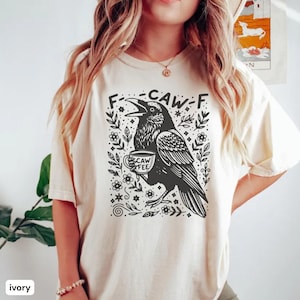 Può includere: T-shirt color avorio con un'illustrazione di un corvo nero che tiene una tazza di caffè. Il corvo è circondato da disegni floreali e di foglie, con la scritta "F-CAW-FEE" sopra. Abbigliamento casual.